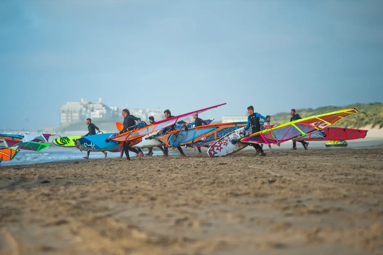 Deelnemers lopen met hun zeilen over het strand